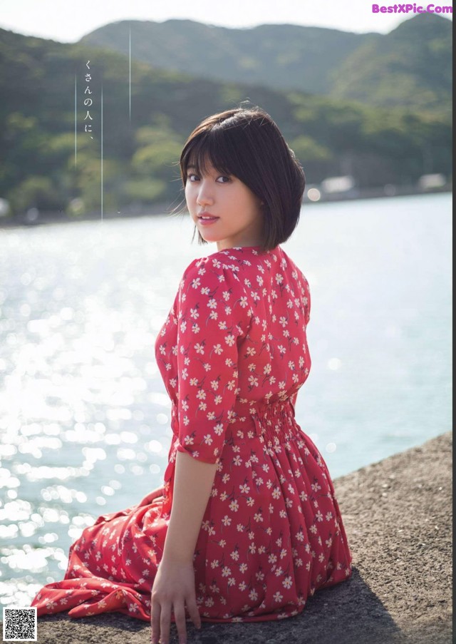 A woman in a red dress sitting on a rock by the water.