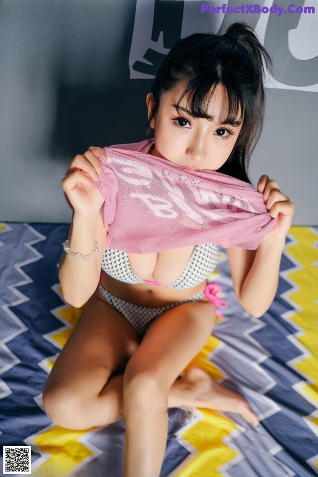 A woman in a bikini sitting on a bed holding a pink scarf.